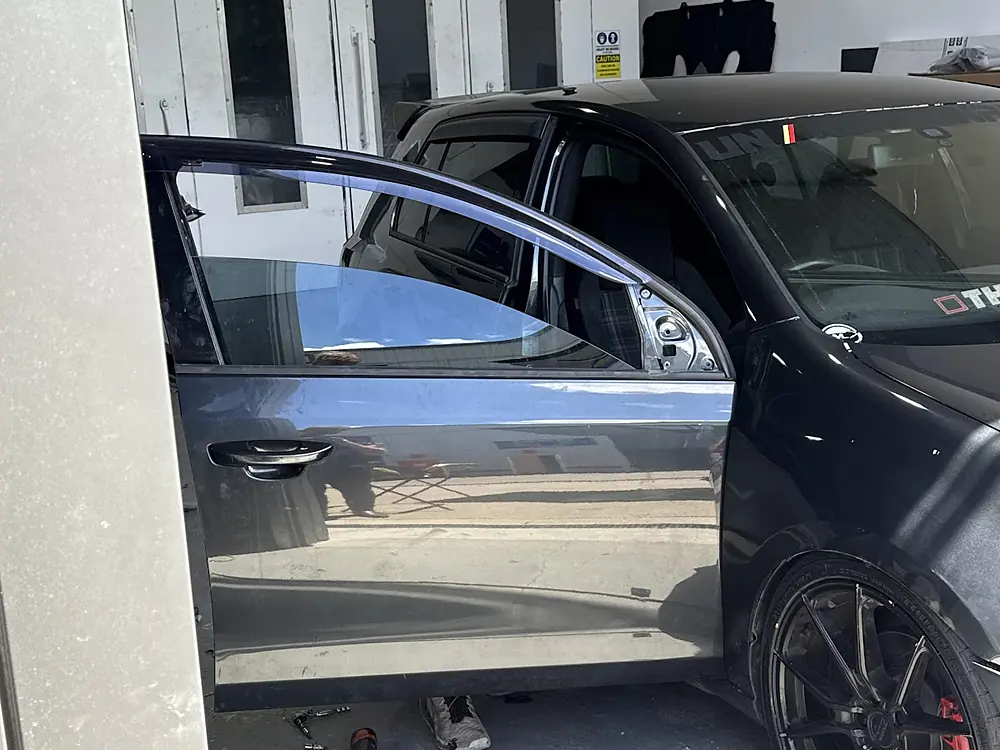 Black hatchback with its front door open in the workshop, beside the spray booth